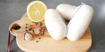 ein Holzbrettchen mit einem Paar Stockwürsten, dekoriert mit Muskatnuss, schwrzen Pfefferkörnern und Zitrone