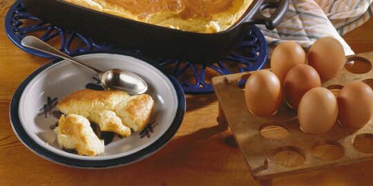 ein Teller mit Mehlkuchen und einem Kaffeelöffel, neben einer Auflaufform und Eiern auf einer Holzhalterung