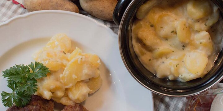 Kartoffelgemüse auf einem Teller mit Petersilie serviert, daneben ein Topf mit Saurem Kartoffelgemüse und rohe Kartoffeln